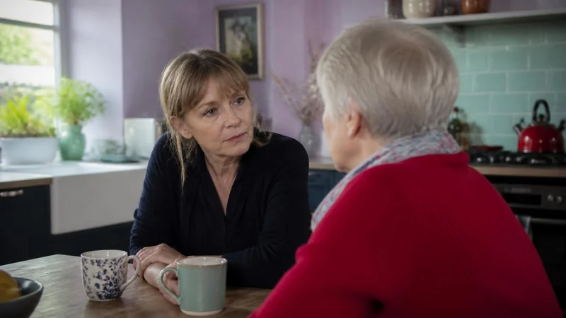 A woman in her fifties sitting at a kitchen table with an older relative, listening, in a lilac and mint kitchen with quiet daylight