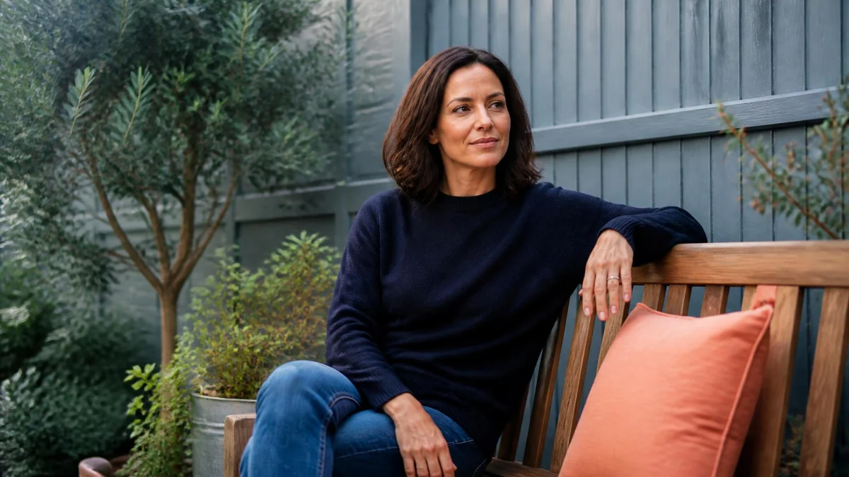 A woman in her forties sitting on a garden bench in a quiet back garden, looking ahead, olive foliage and apricot cushions