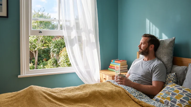 a thirty something male, not wearing a hat, is ill in bed, he is wearing a t shirt,  with the sun shi=ning outside his bedroom window, his sash window is open an d blowing his chiffon curtain gentl in the breeze