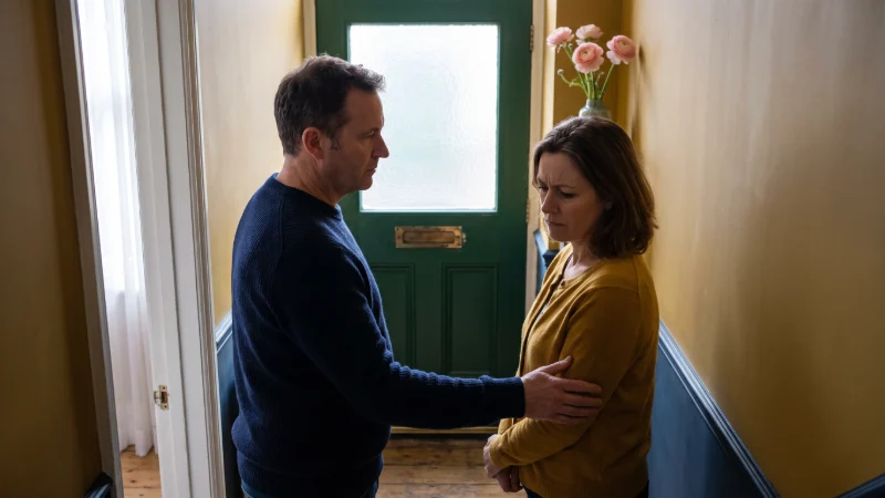Two adult siblings talking SOMBERLY NOT SMILING Standing  in the hallway of a terraced house,  the closed front door is behind them