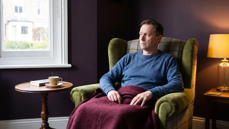 A man in his fifties, looking ill and grey,  resting in an plaid armchair by a window, with a small side table, burgundy blanket across his knees